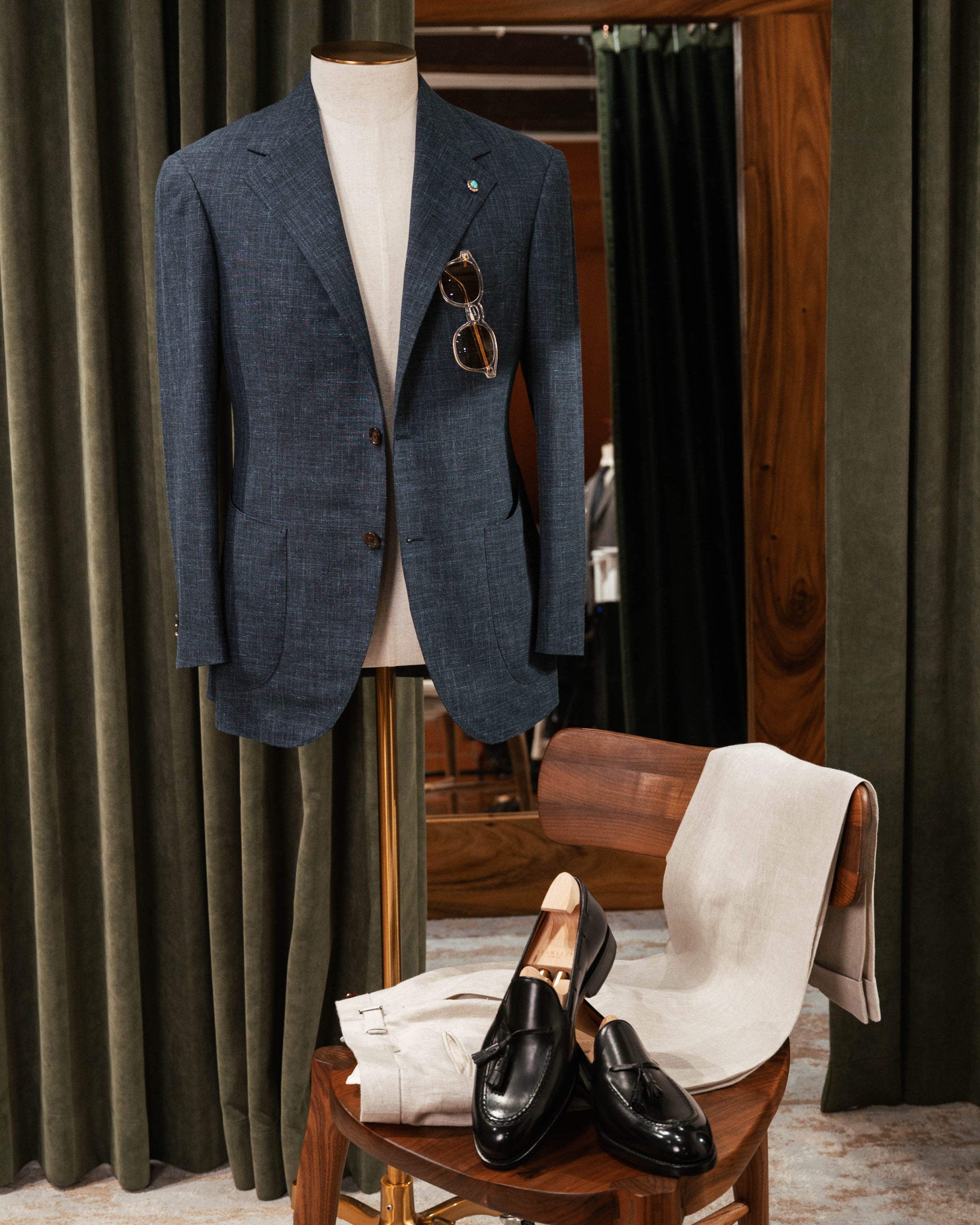 Blue suit jacket on a mannequin with sunglasses, black shoes on a chair, and green curtains in the background.