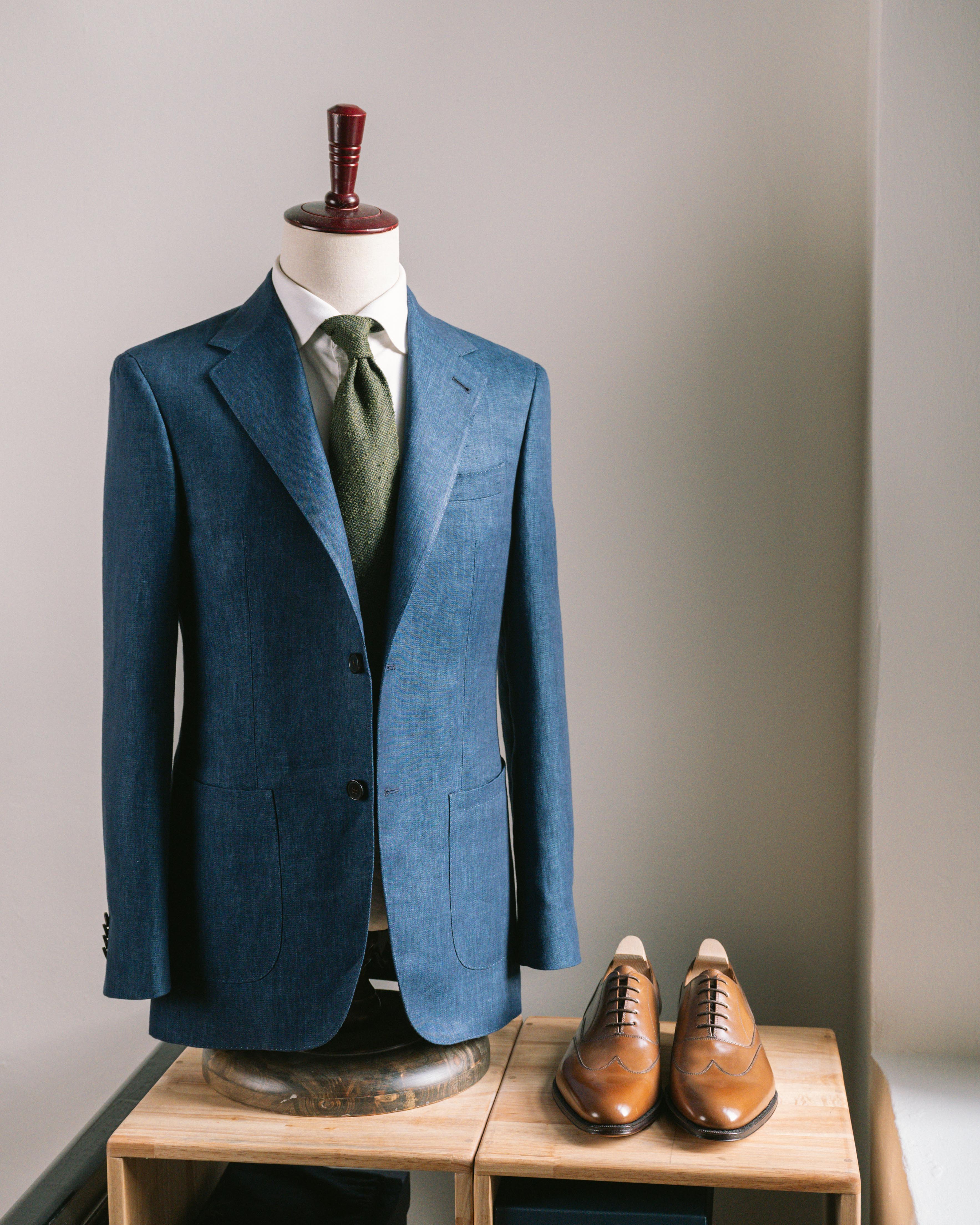 Blue suit jacket with a green tie on a mannequin, paired with brown shoes on a wooden stand against a neutral background.